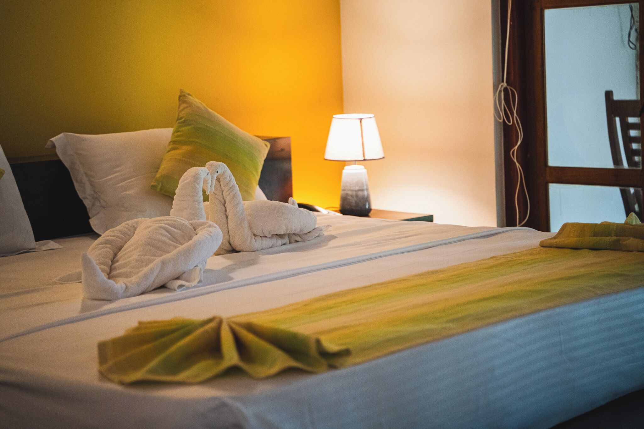 A hotel room with a neatly made bed, a lamp on the nightstand, and a window with a view.