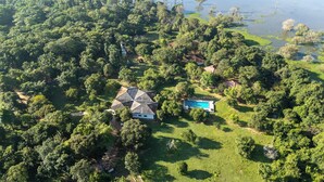An aerial view of a property with a large house featuring a unique geometric roof, surrounded by dense greenery and a swimming pool. - Pothana Village (Sigiriya)