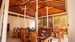 A spacious dining area with wooden tables and chairs, large windows, and ceiling fans. - Pothana Village (Sigiriya)