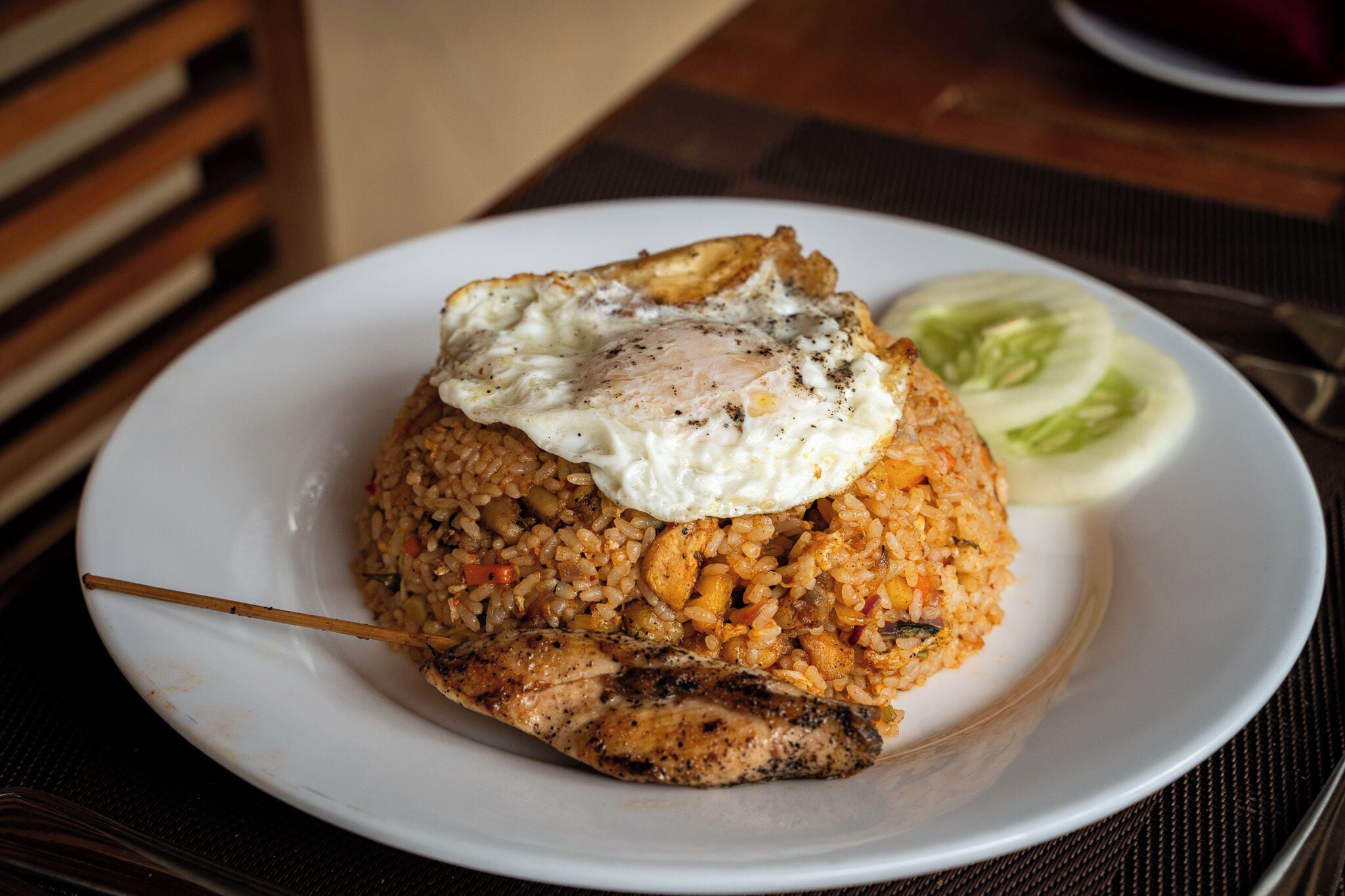 Une assiette de riz frit, garnie d’un œuf au plat et d’un morceau de poulet grillé, le tout présenté sur une assiette blanche.