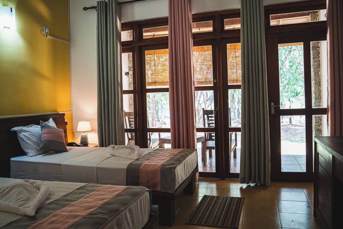 Quarto de hotel com duas camas, piso de madeira, escrivaninha com luminária e janelas amplas com cortinas.
