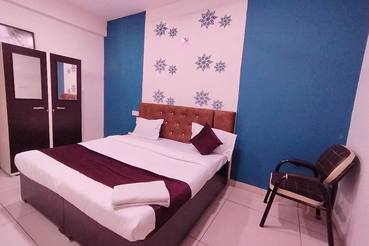 A neatly made bed with a brown headboard, white and maroon pillows, and a white blanket. There is a chair with a dark cushion and a blue wall with floral patterns.