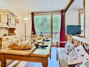A cozy dining area with a wooden table set for breakfast, a television on a small table, and a balcony with a view of trees.