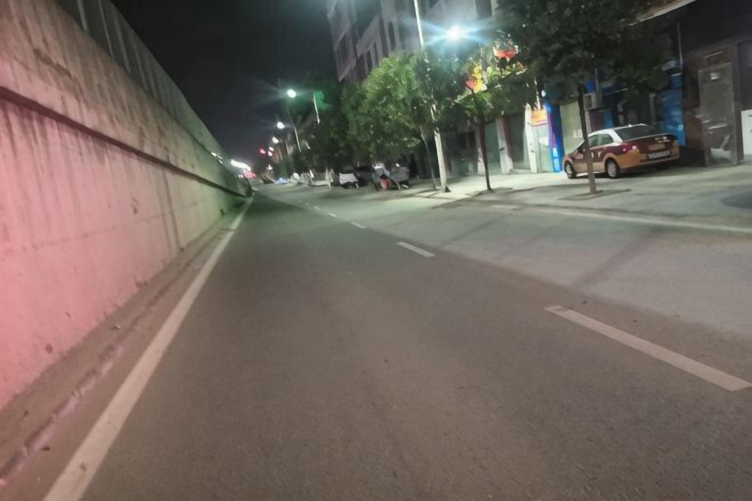 Une vue nocturne d’une rue, avec une route, un mur rose et une voiture garée.