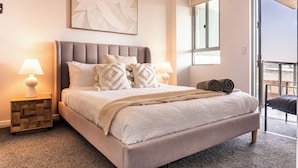 A neatly arranged bedroom with a large bed, two bedside lamps, a wooden nightstand, a window with a view, and a balcony door.