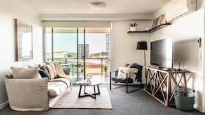 A modern living room with a sofa, coffee table, and TV stand. There is a balcony with a view of buildings and a cityscape.