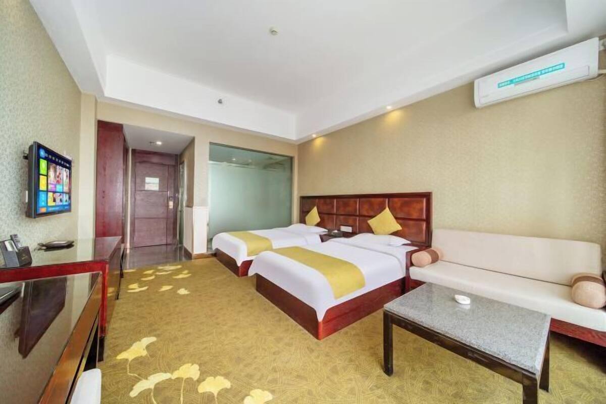 A hotel room with two beds, a sofa, a small table, a TV, and a bathroom visible through a glass door.
