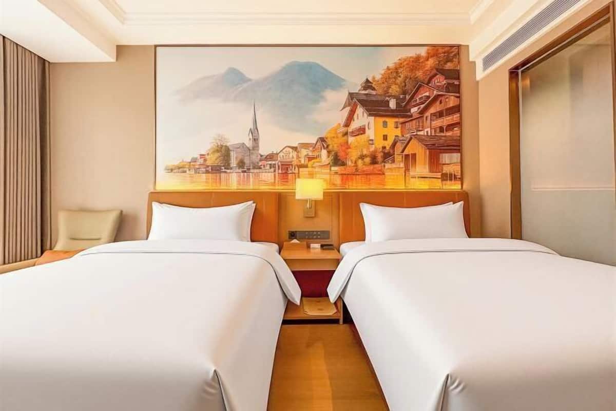 A hotel room with two beds, a painting of a town, a nightstand, and a lamp.