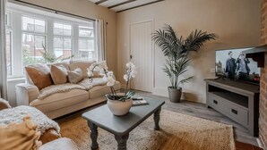 Un salón con un sofá color beige, una mesa de centro con una planta, un televisor sobre un soporte de madera y ventanas amplias con cortinas.