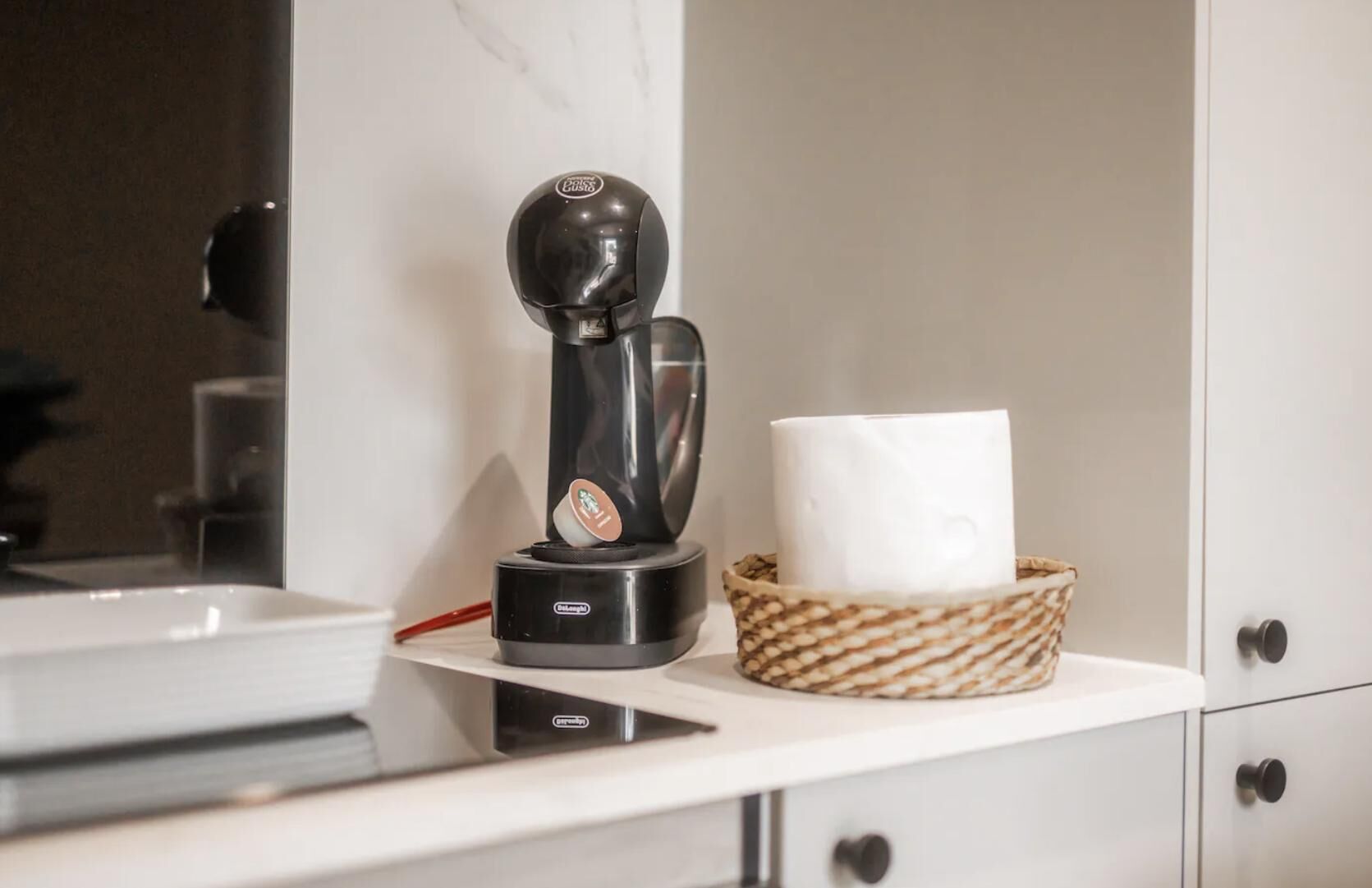 Une machine à café posée sur un plan de travail blanc, une tasse blanche dans un panier en osier et un meuble blanc avec un tiroir.
