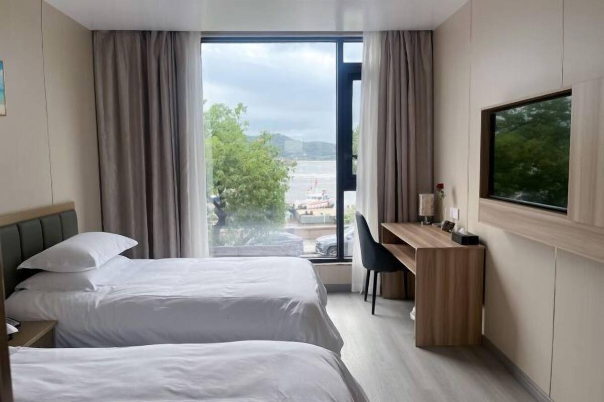 A hotel room with two beds, a desk, a chair, and a large window with a view of the outdoors.