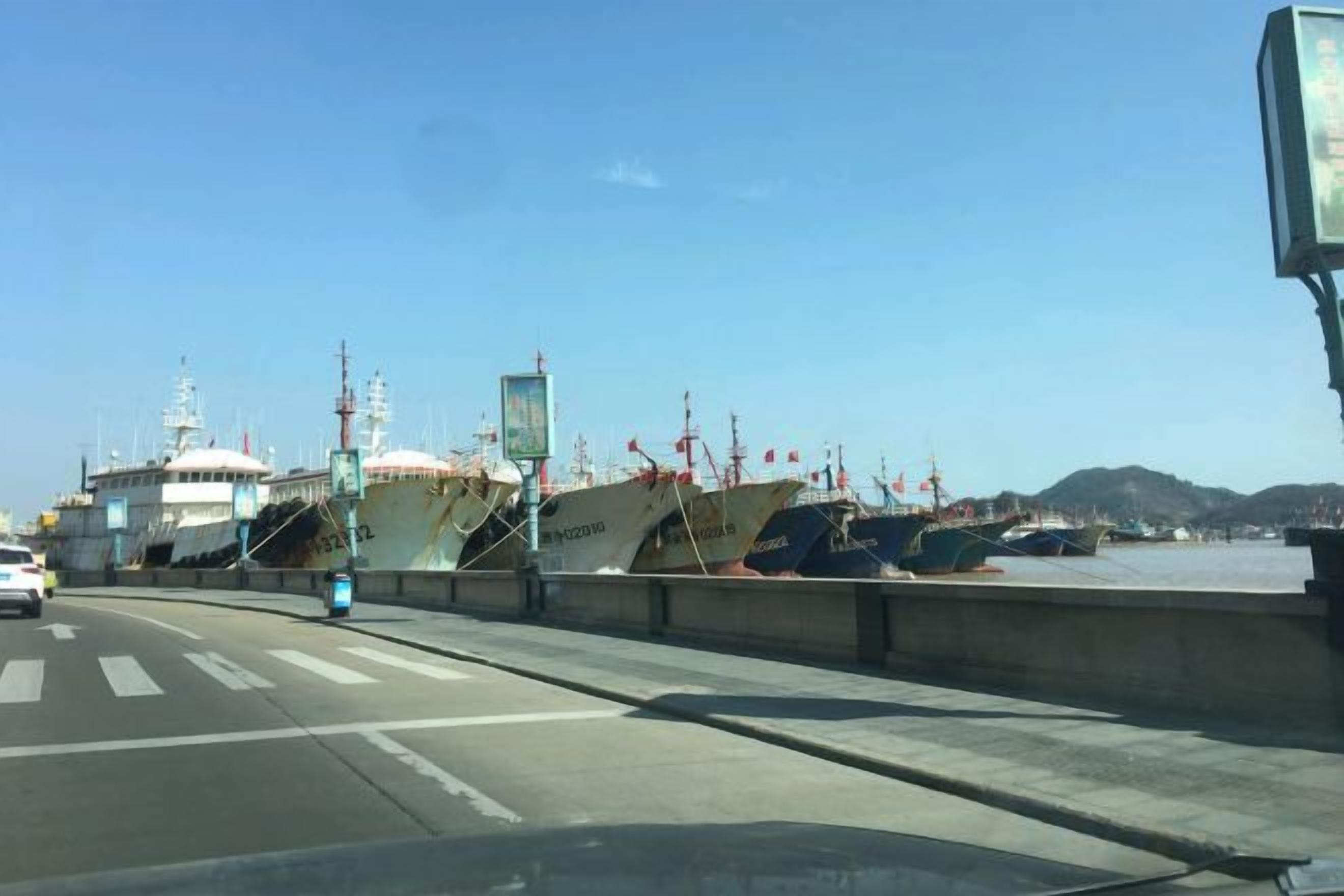 A coastal road with a bridge, boats docked, and a clear sky.