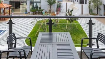 A balcony with a table and chairs, overlooking a checkered pavement and a green lawn.