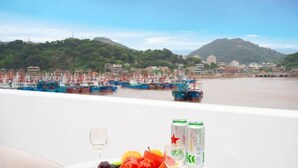 A table with a fruit platter, wine glass, and two cans of water, overlooking a harbor with boats and a mountainous backdrop. - TongQianWan Hotel (Xiangshan)