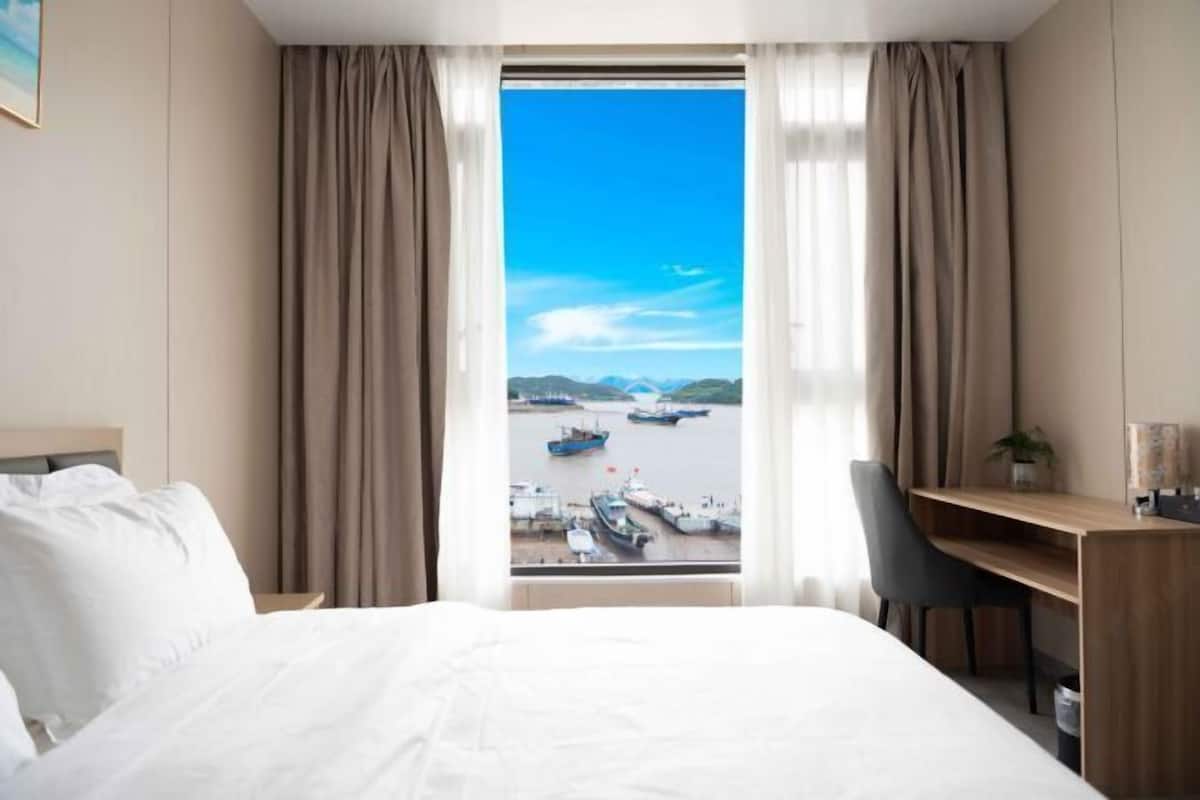A hotel room with a bed, a desk, and a view of boats at a harbor.