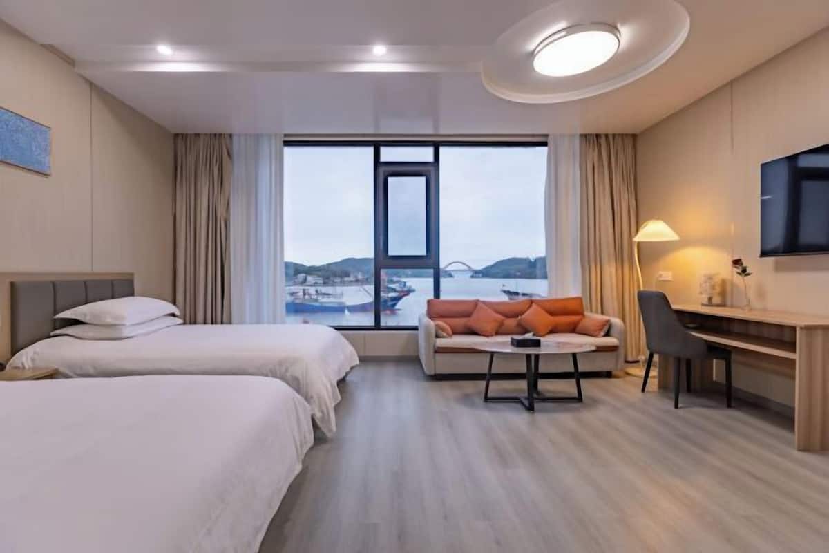 A hotel room with two beds, a sofa, a desk, a chair, a TV, and a large window with a view of a bridge and boats.