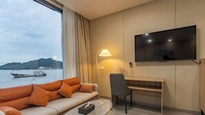 A hotel room with a sofa, a desk, a TV, and a view of a body of water. - TongQianWan Hotel (Xiangshan)