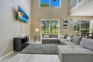 Een ruime woonkamer met een grote grijze modulaire bank, een flatscreen televisie aan de muur en een groot raam met uitzicht op de omgeving.