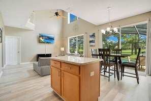 A spacious kitchen with a large island, a dining area with a table and chairs, a ceiling fan, and a view of the outdoors through large windows.