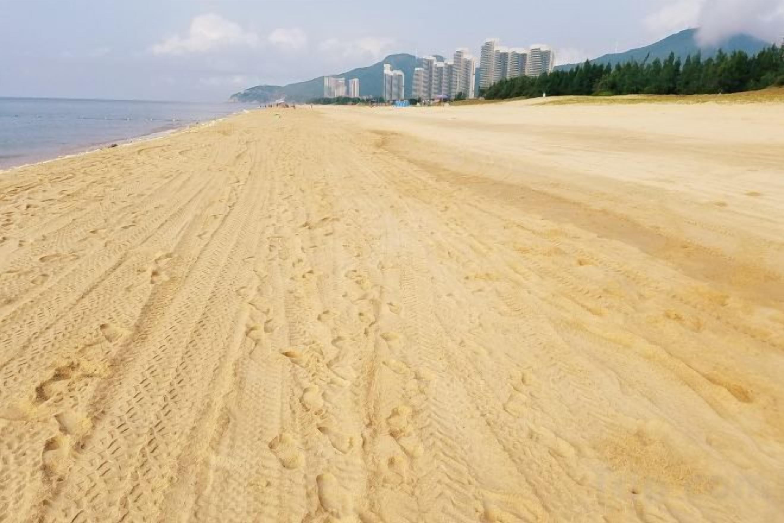 Een uitgestrekt zandstrand met voetafdrukken, een heldere lucht en gebouwen op de achtergrond.
