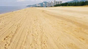 Una amplia playa de arena con huellas, cielo despejado y edificios al fondo.