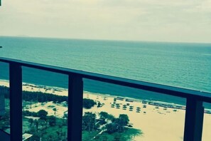 Blick auf den Ozean von einem Balkon mit Geländer, ein Strand mit Bäumen und Sonnenschirmen in der Ferne und ein klarer Himmel.