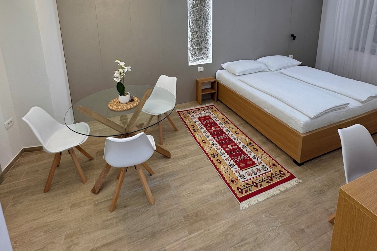 A modern bedroom with a bed, a glass dining table with chairs, a decorative rug, and a ceiling light fixture.