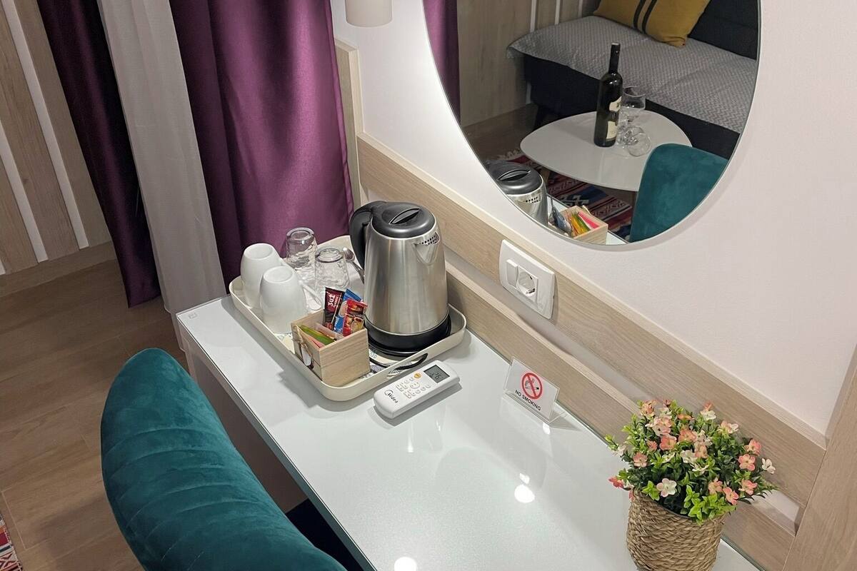 A modern hotel room with a white desk, teal armchairs, a coffee maker, a digital scale, a small potted plant, and a mirror reflecting the room.