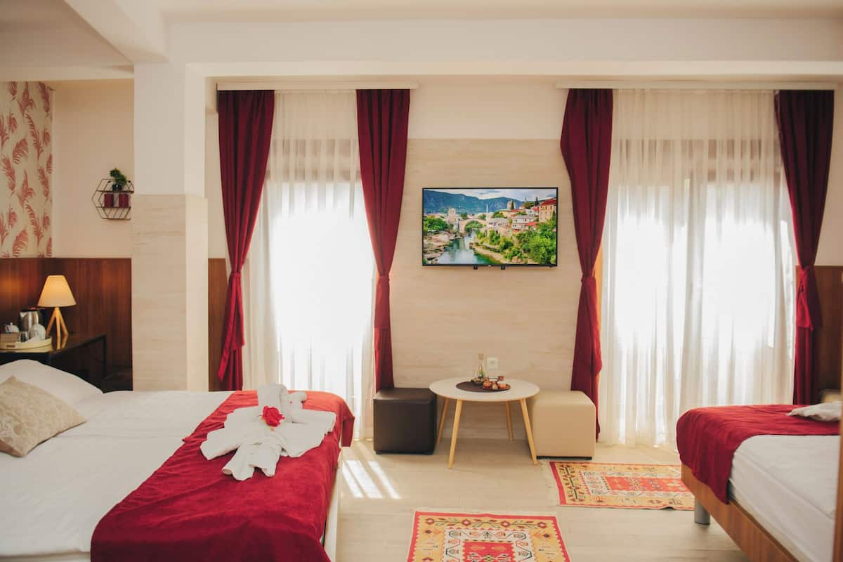 A hotel room with two beds, a small table with a vase, a wall-mounted TV, and a window with curtains.