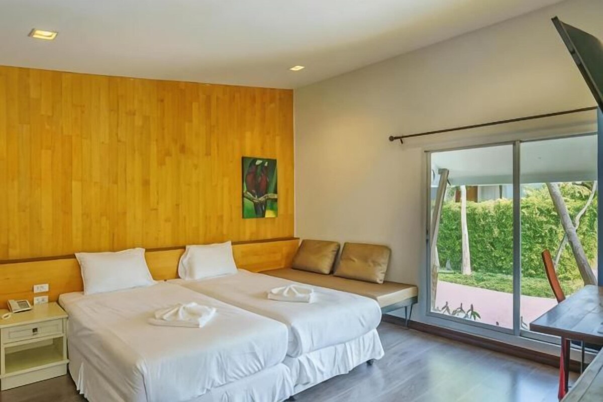 A hotel room with two beds, a wooden wall, a large window, and a view of greenery.