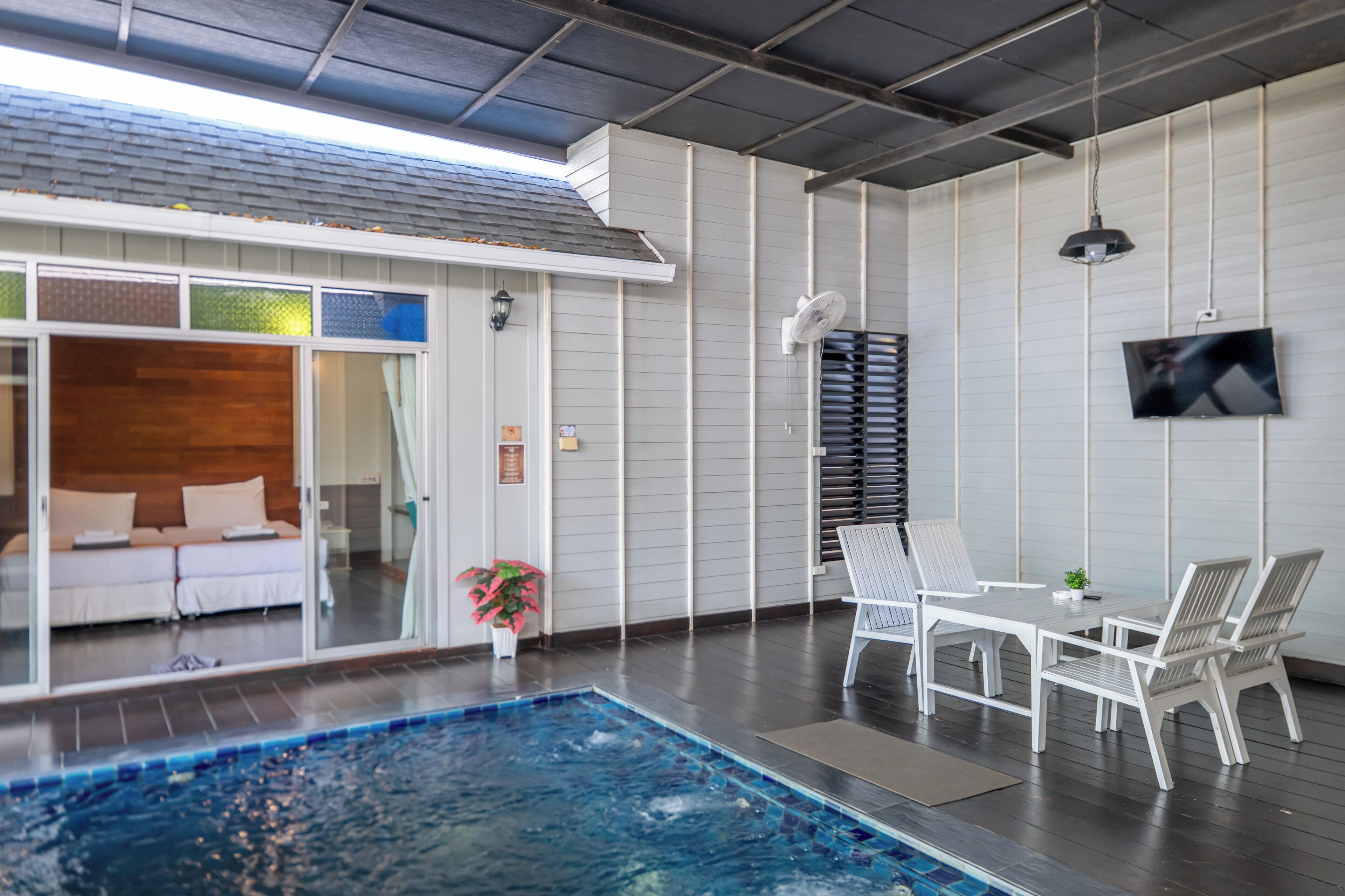 A modern indoor pool area with a glass door leading to a bedroom, a dining area with white furniture, and a flat-screen TV mounted on the wall.