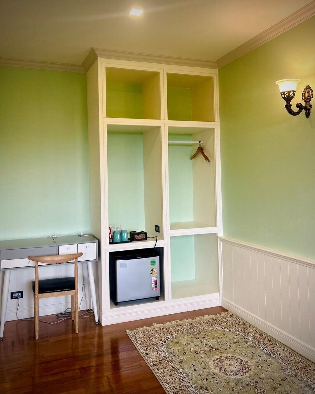 A room with a desk, a cabinet with a hanging clothes rack, a small refrigerator, and a patterned rug.