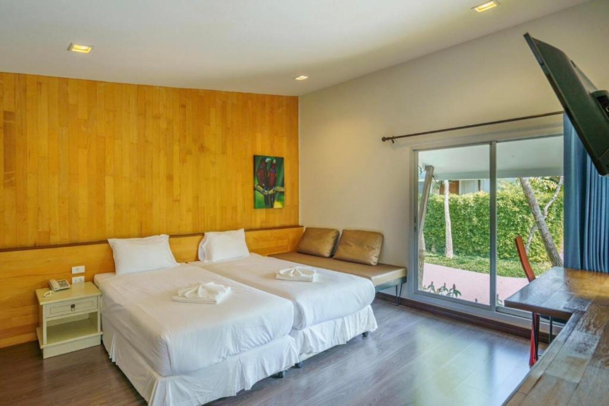 A hotel room with two beds, a wooden wall, a painting, a bench, and a view of greenery through large windows.