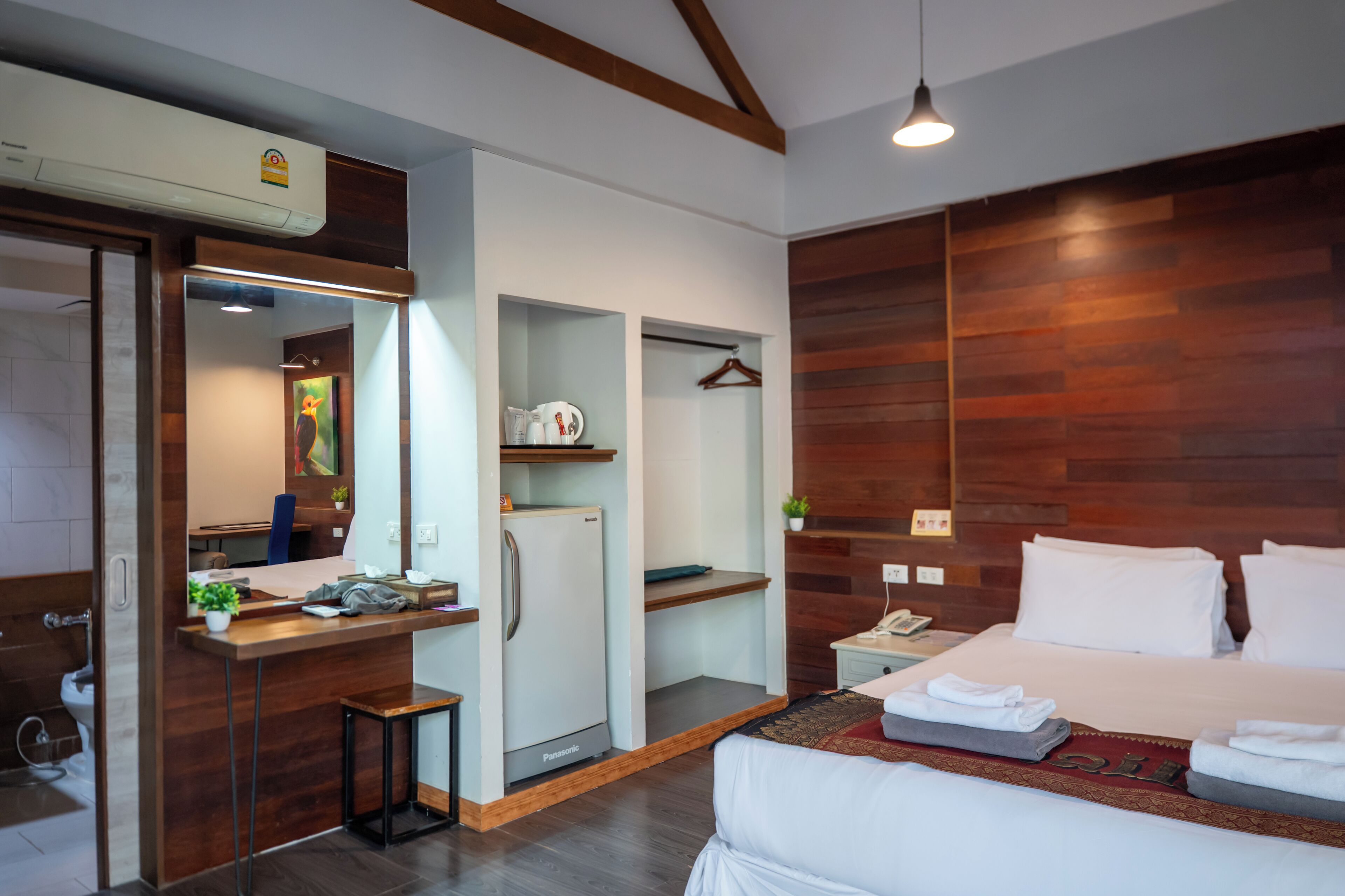 A modern hotel room with a wooden accent wall, a bed with white linens, a desk with a computer, a mini-fridge, and a small seating area.