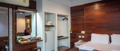 A modern hotel room with a wooden accent wall, a bed with white linens, a desk with a computer, a mini-fridge, and a small seating area.