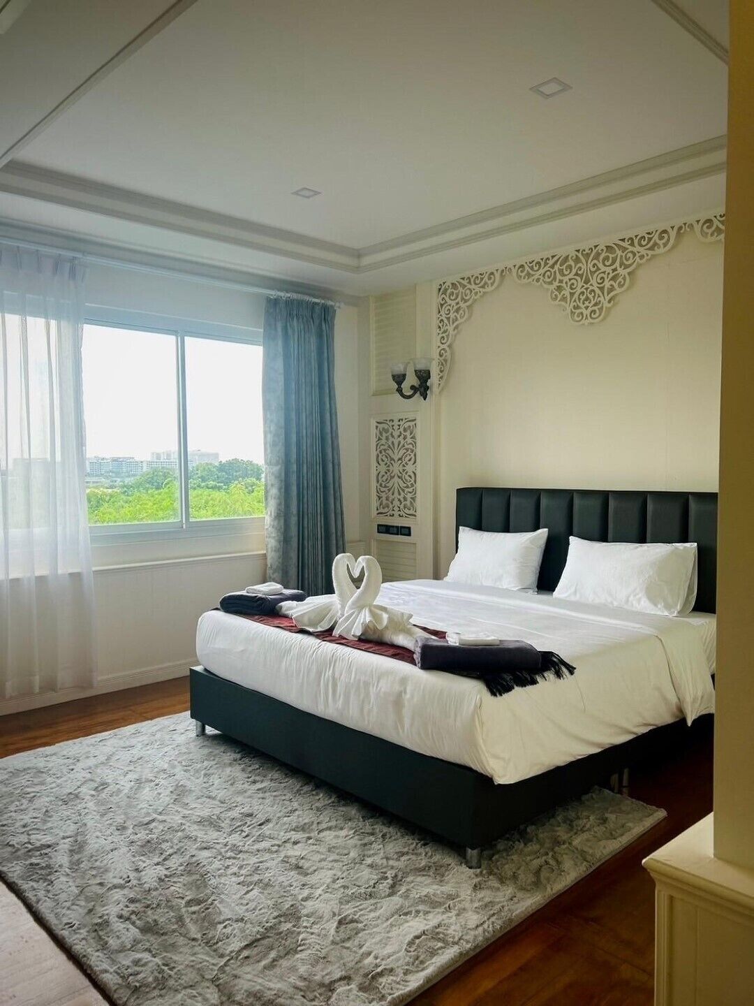 A hotel room with a large bed, a view of greenery through the window, a decorative wall piece, and a fluffy rug.