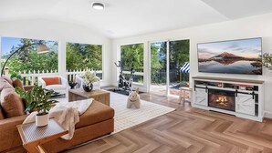 Ein geräumiges Wohnzimmer mit Kamin, großem Fernseher und Blick auf eine Terrasse und Bäume.
