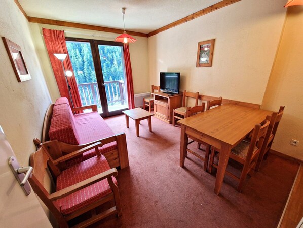 Una habitación acogedora con una mesa de comedor de madera, sillas, un sofá, un televisor y un balcón con cortinas rojas.
