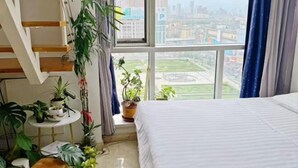 A bedroom with a bed, a window with a city view, a staircase, and several potted plants. - Kaixuan Shidai Family Apartment (Dalian)