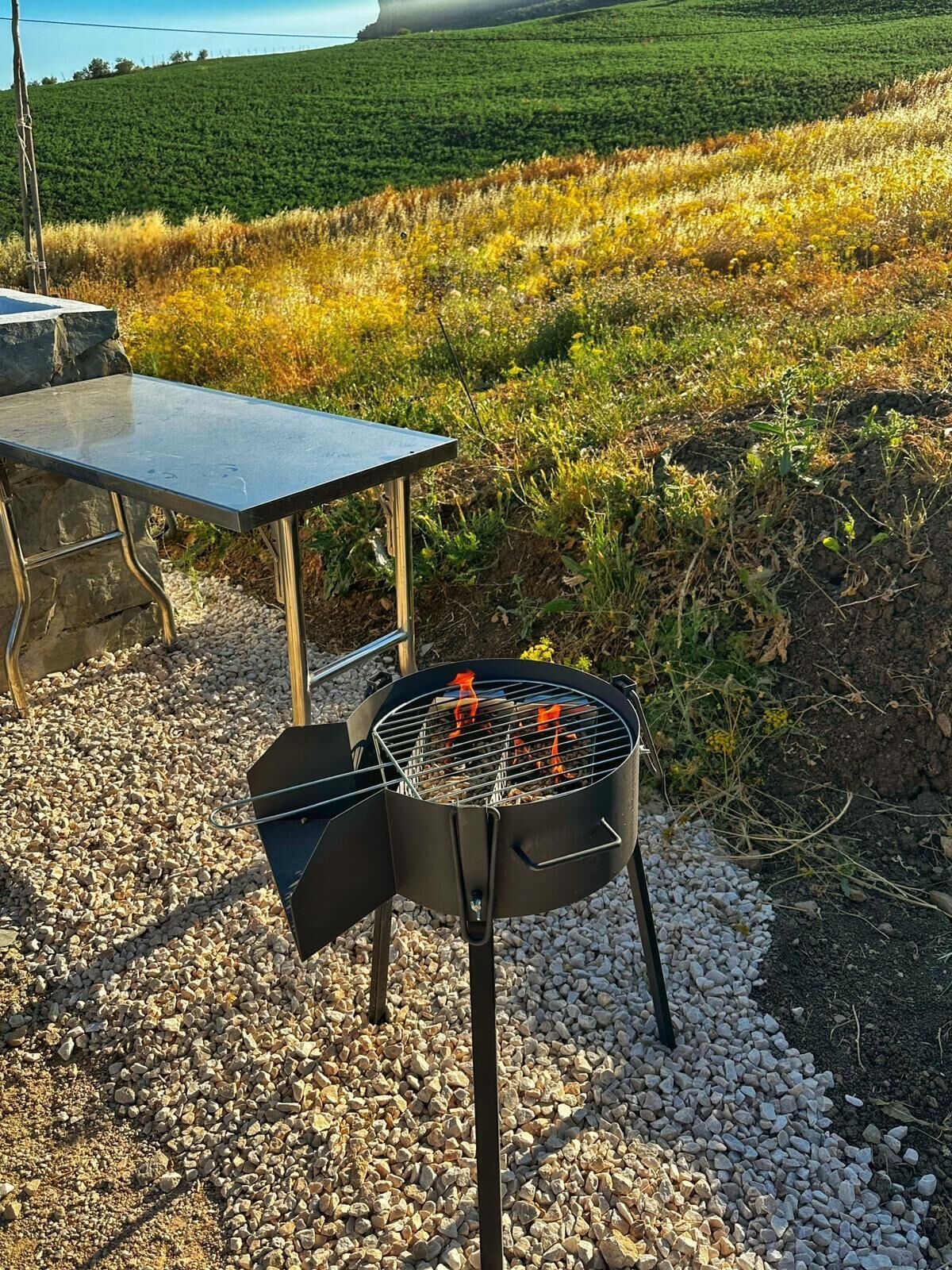 Una parrilla al aire libre con una mesa y sillas, ubicada sobre un terreno de grava con un campo verde de fondo.
