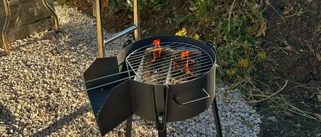 Una parrilla al aire libre con una mesa y sillas, ubicada sobre un terreno de grava con un campo verde de fondo.