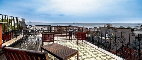 A rooftop terrace with wooden furniture and a patterned floor, overlooking a body of water and distant cityscape.