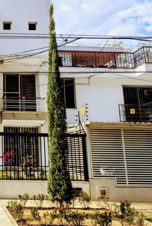 Un immeuble résidentiel de deux étages doté d’un balcon, d’un grand arbre à feuilles persistantes et d’une entrée fermée par un portail.