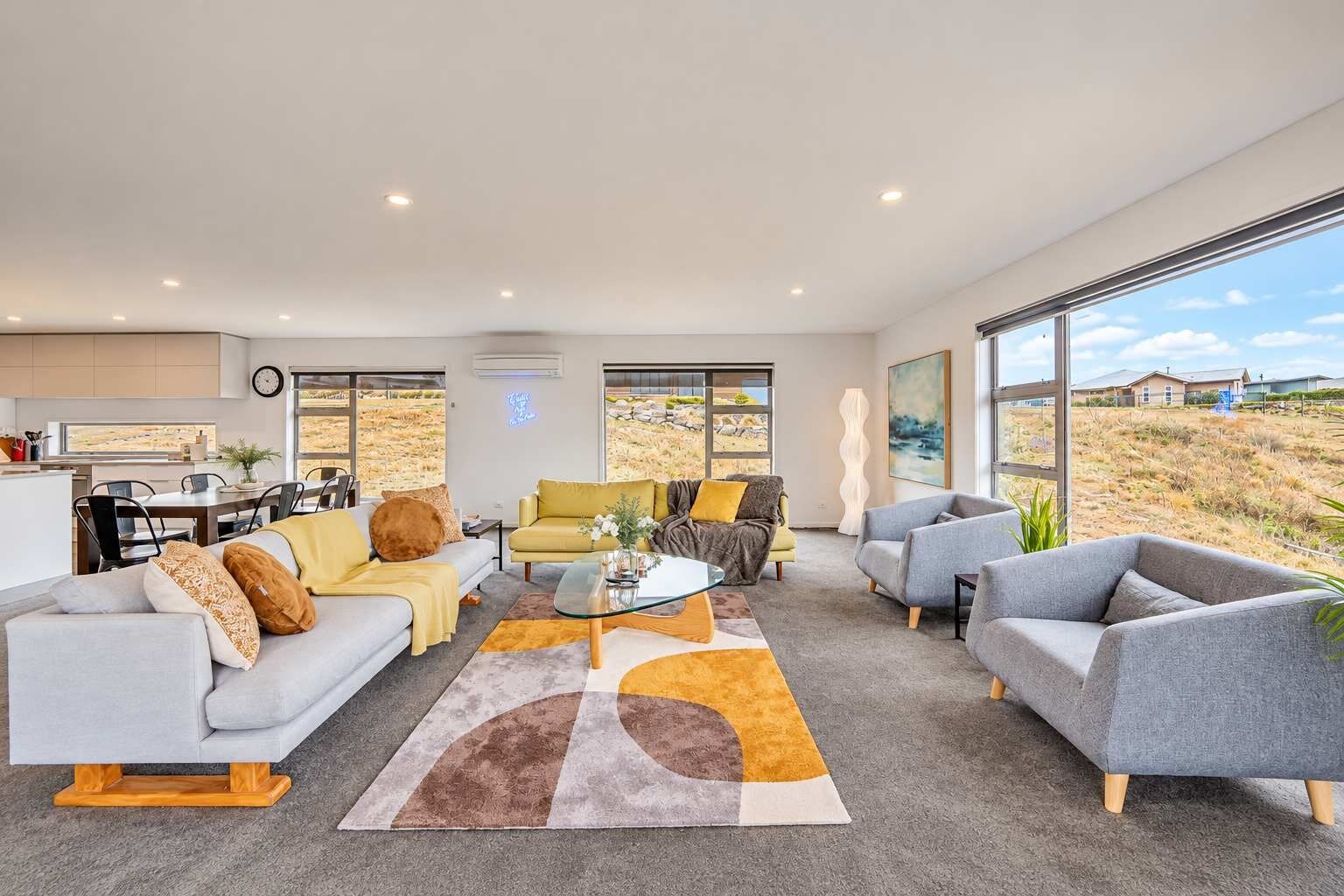 A modern living room with a sofa, armchairs, and a coffee table, large windows with a view of the landscape, and a kitchen area with a dining table.