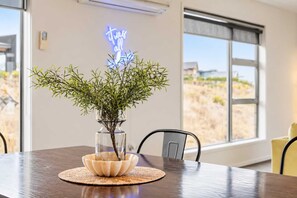 A dining table with a clear glass vase containing green foliage, a woven placemat, and a neon sign on the wall.