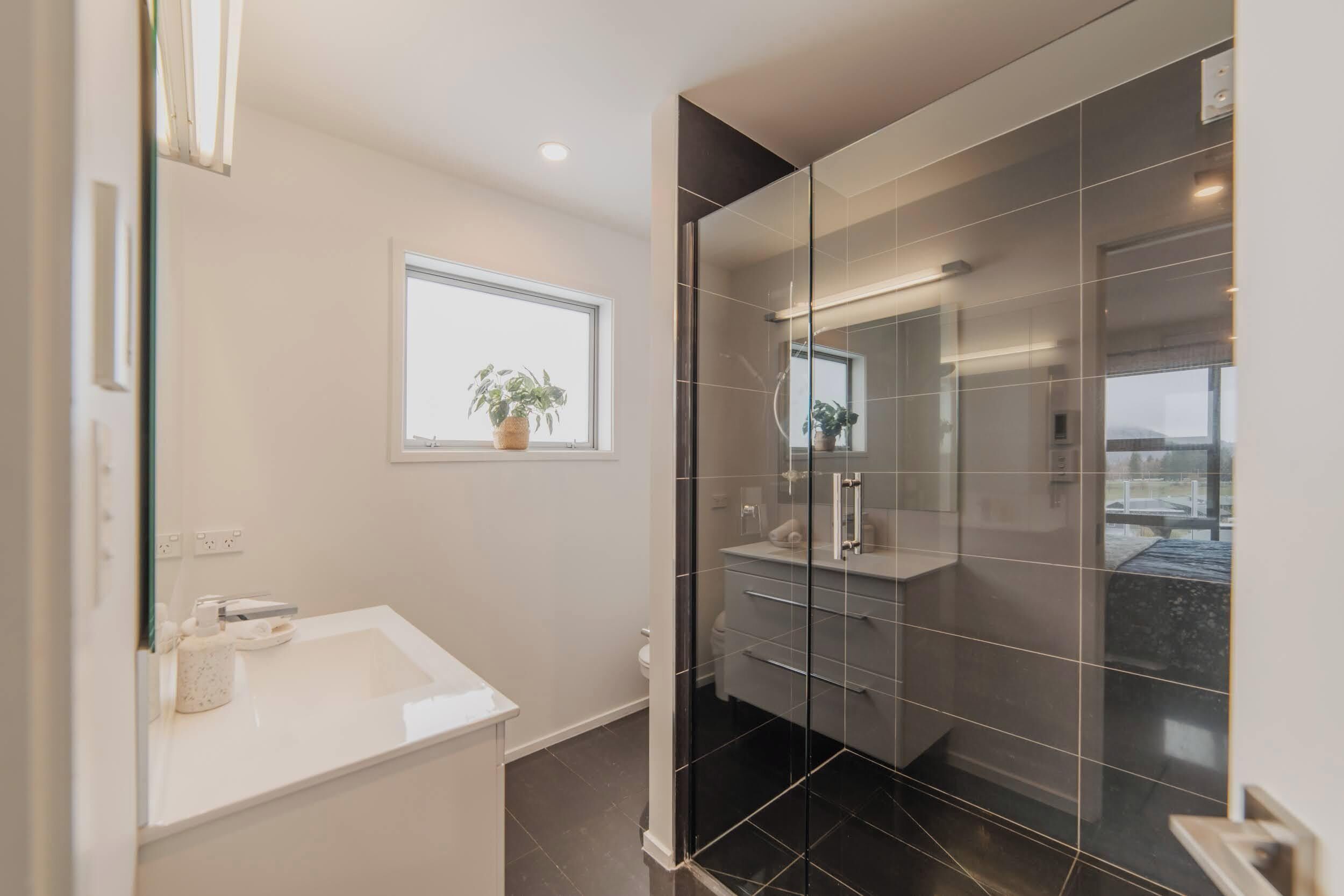 A modern bathroom with a glass shower enclosure, a white sink, and a window with a plant.