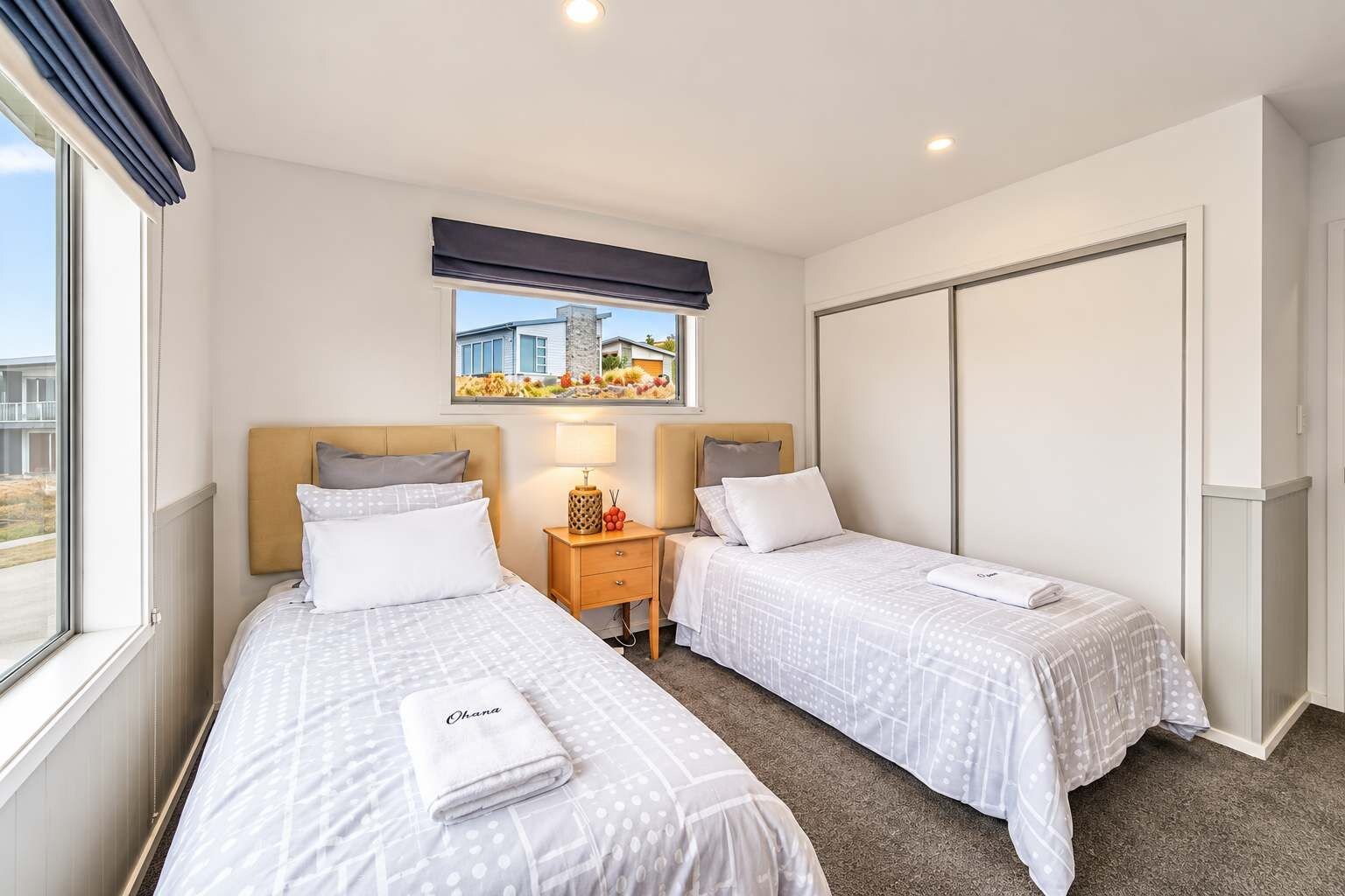 A hotel room with two beds, a window with a view, a bedside table with a lamp, and a wardrobe.