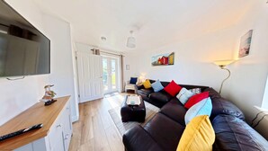 A living room with a black leather sofa, a wooden coffee table, a TV, and a door leading to an outdoor area. - Stunning Coastal Cottage - Sleeping 6 in 3 Bedrooms - Pool and Beach Access Included. (Filey)