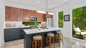 A modern kitchen with dark cabinets, a large island, and bar stools. - Modern Treehouse Retreat, Cold Plunge (Austin)