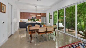 A modern kitchen with wooden cabinets, a central island, and a dining area with a large window overlooking a lush garden. - Modern Treehouse Retreat, Cold Plunge (Austin)